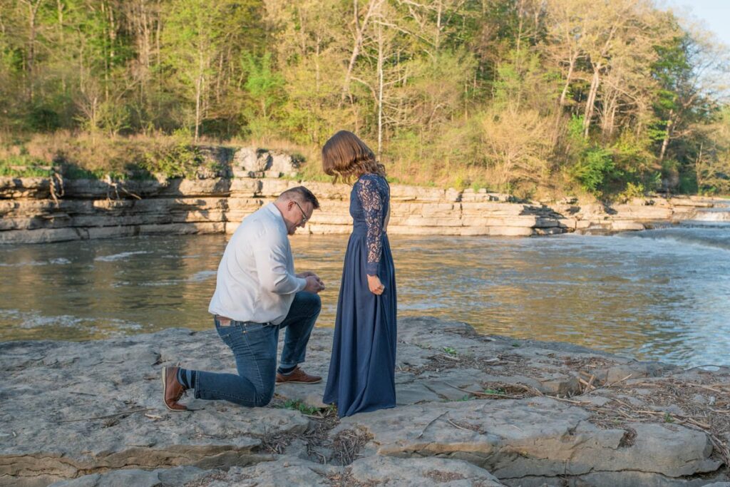 Man gets down on one knee to propose to woman on a rock in front of a waterfall.