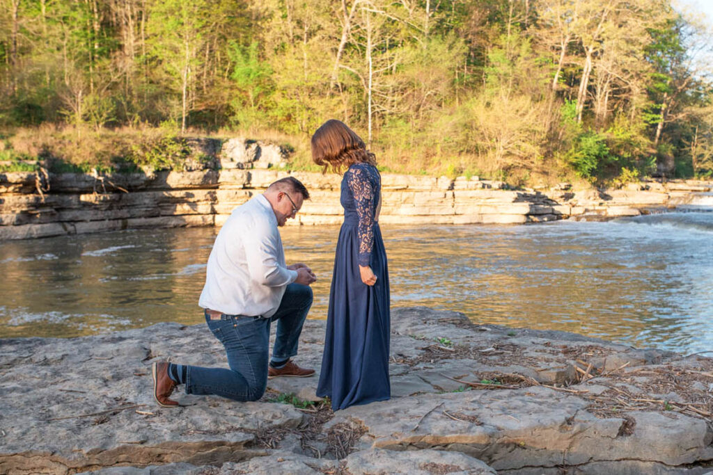 Man gets down on one knee to propose to woman on a rock in front of a waterfall.