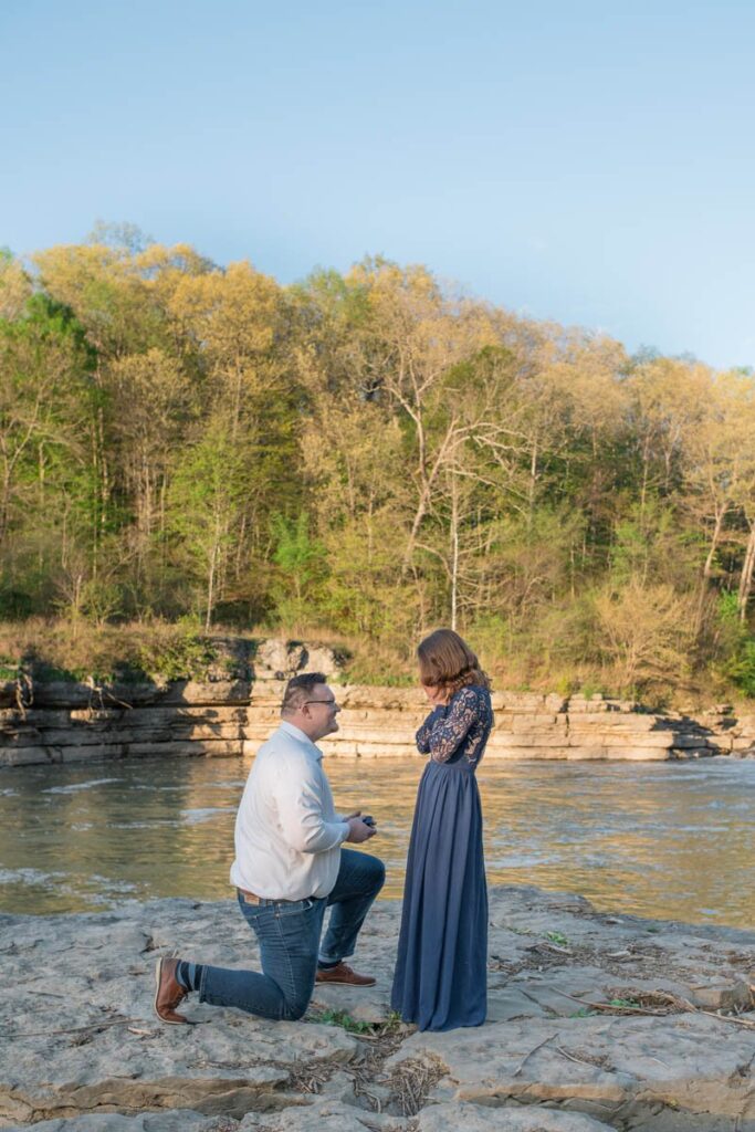 Proposal photographer captures man proposing to woman in front of waterfall while woman has her hands on her face surprised.