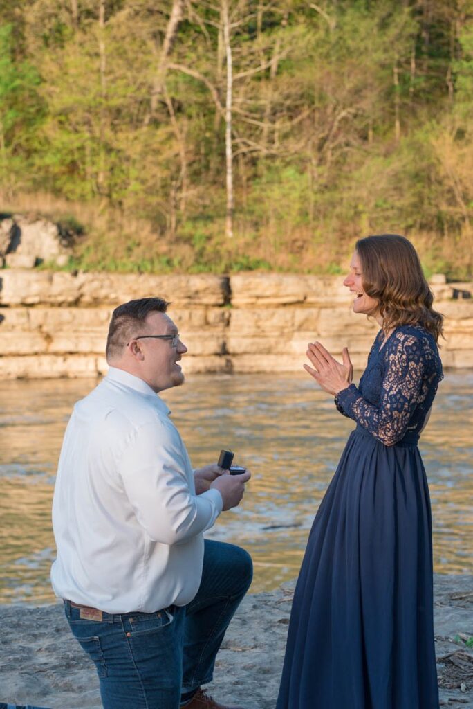 Proposal photographer captures surprise engagement with man on one knee holding ring box towards smiling woman.