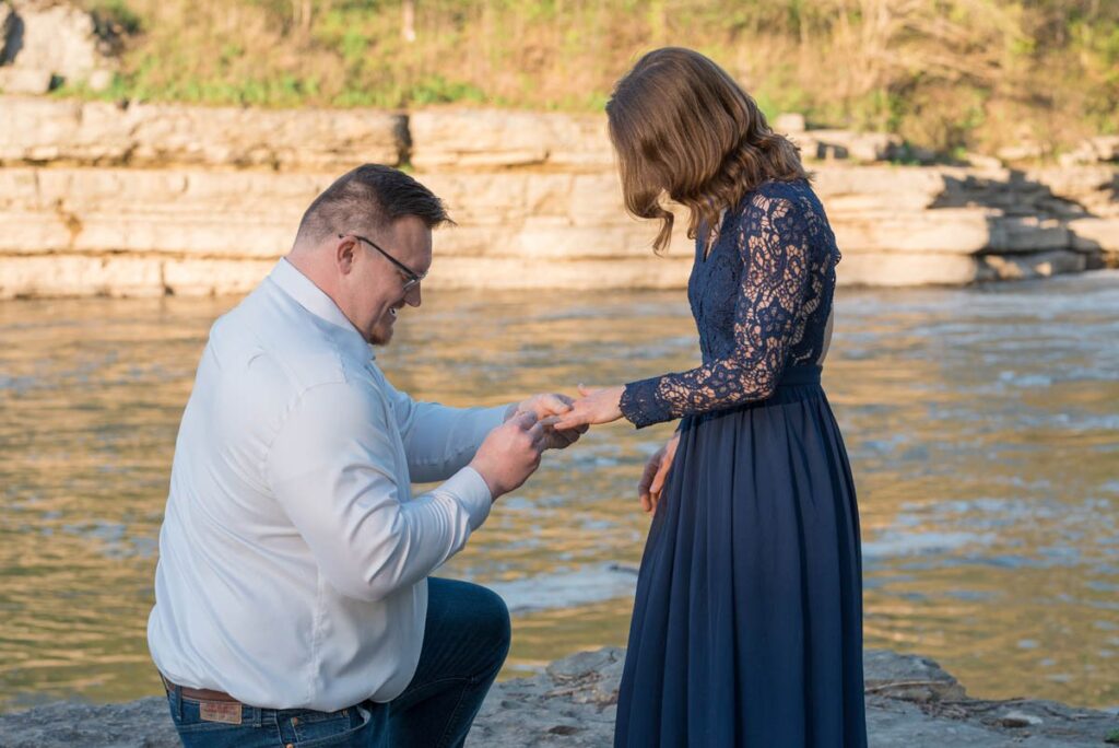 Man is down on one kneed and puts engagement ring on her finger in front of waterfall.