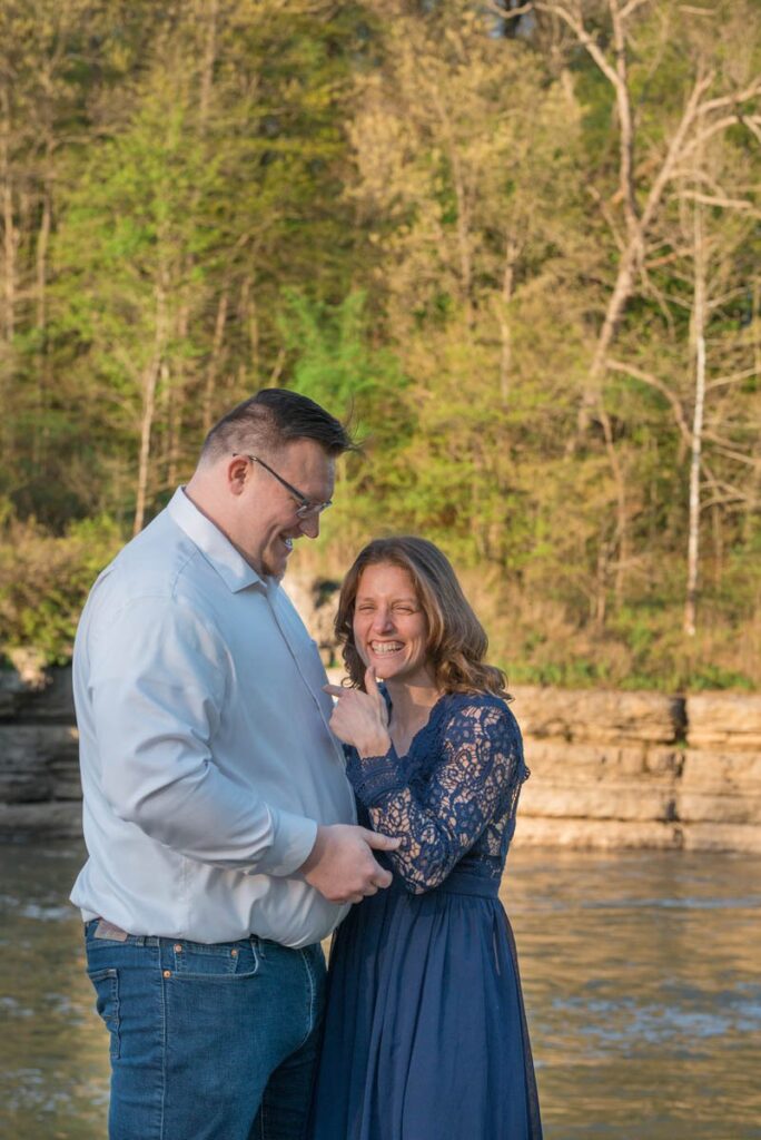 Couple hugs while woman laughs, looks at proposal photographer, and points to man.