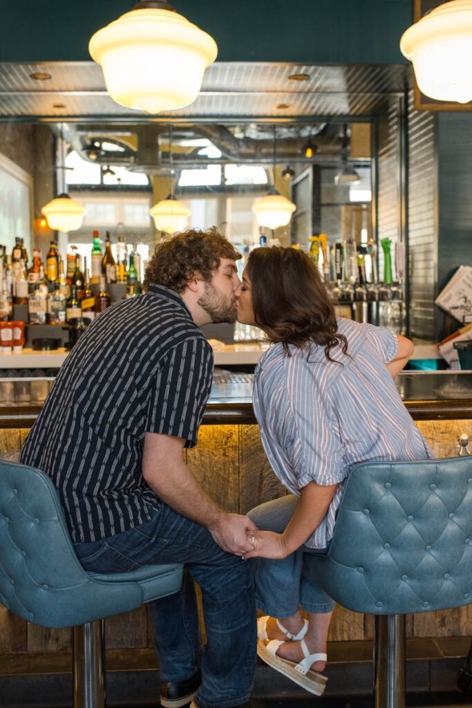 Couple kisses while sitting in barstools in front of a bar.
