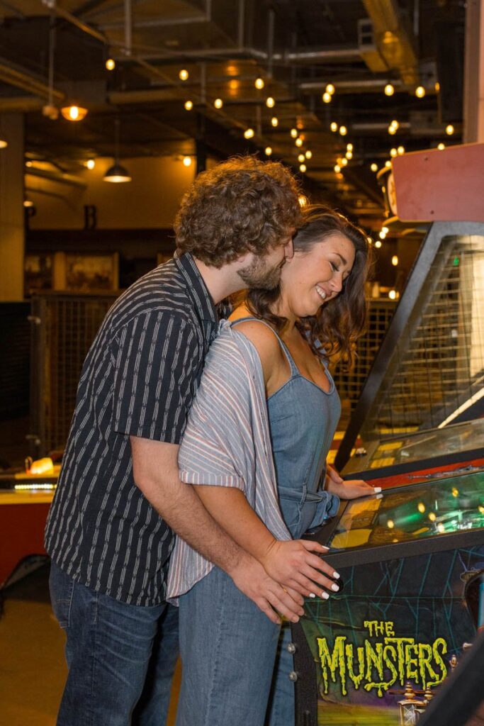 Man kisses woman on the side of her face as they play pinball.