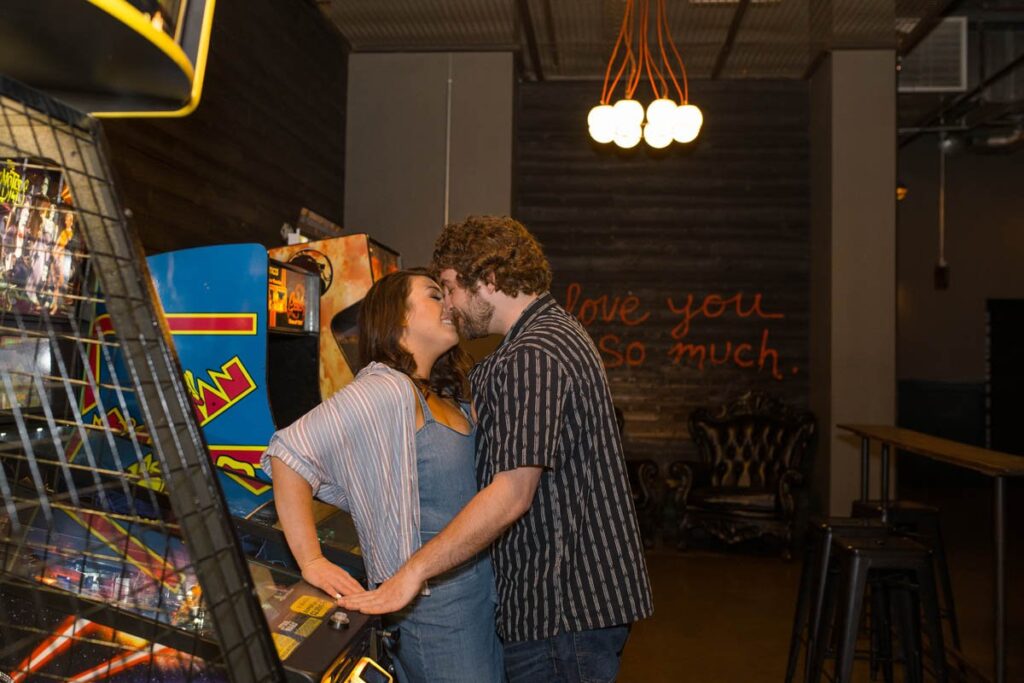 Man leans woman against an arcade game to kiss her at Punch Bowl Social for their Indianapolis engagement photos.