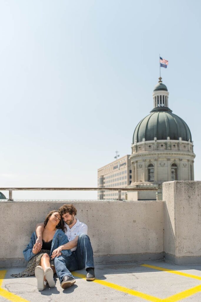 Couple sits on a rooftop parking garage with arms around each other and eyes closed.