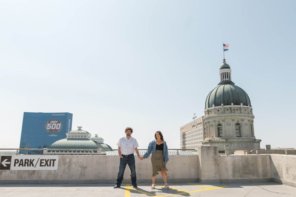 Couple stands on a rooftop parking garage in Indianapolis with the State Capital Building behind them during Indianapolis engagement photos..