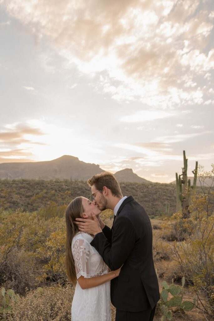 Bride and groom kiss in the desert at sunset in front of mountains.