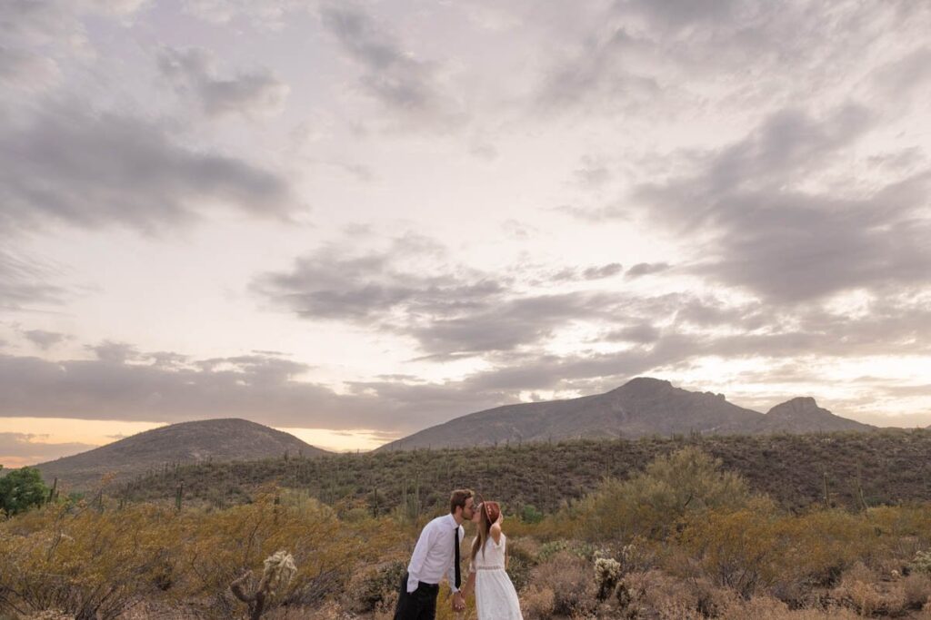 Bride and groom kiss in front of mountains while bride holds onto hat.
