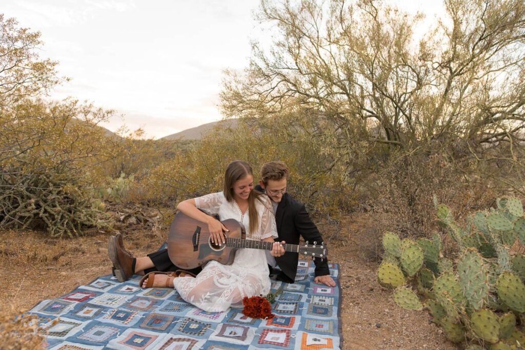 Couple sits on a blanket in the desert playing the guitar together at sunset.