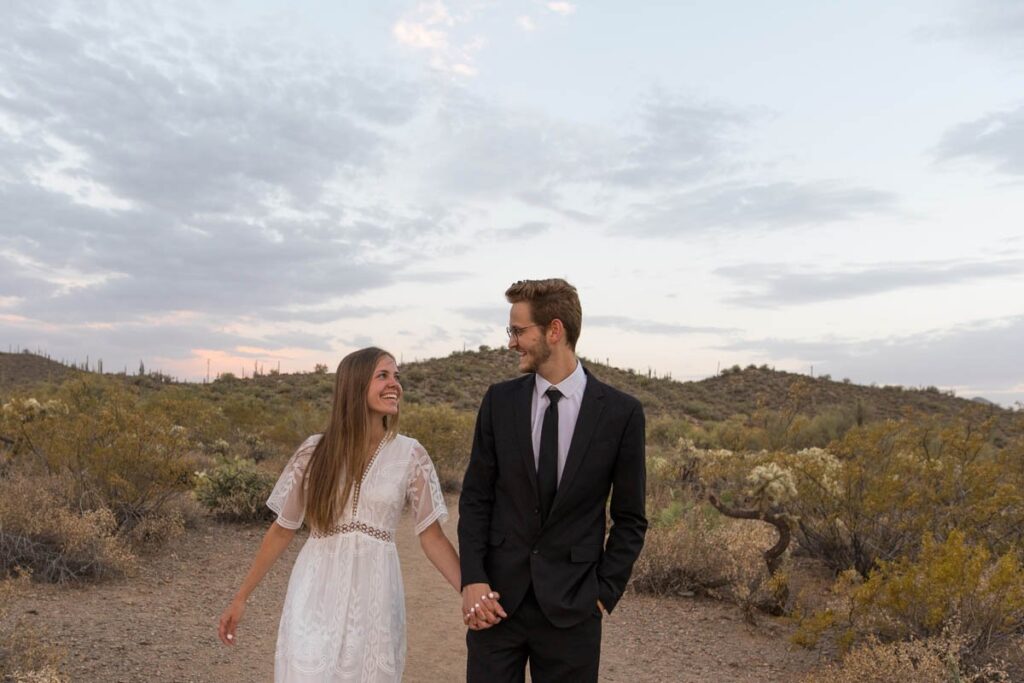 Bride and groom smile at one another while walking in the desert at sunset after eloping in Arizona.