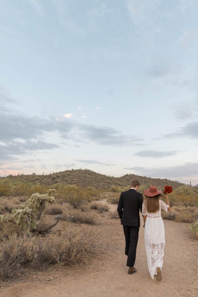 Bride and groom walking on a trail in the desert while bride holds onto hat.