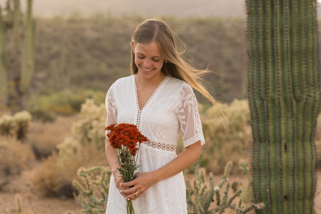 Bride smiles and looks down at her flowers while standing next to a cactus in the desert.
