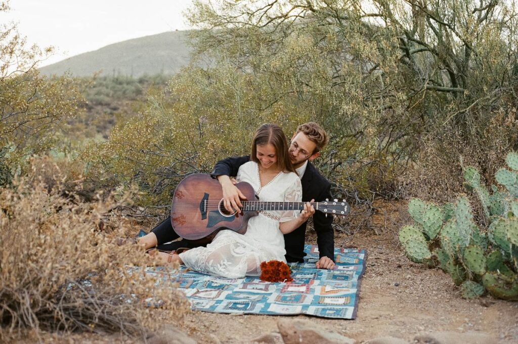 Couple plays guitar together on a blanket in the desert by cacti.