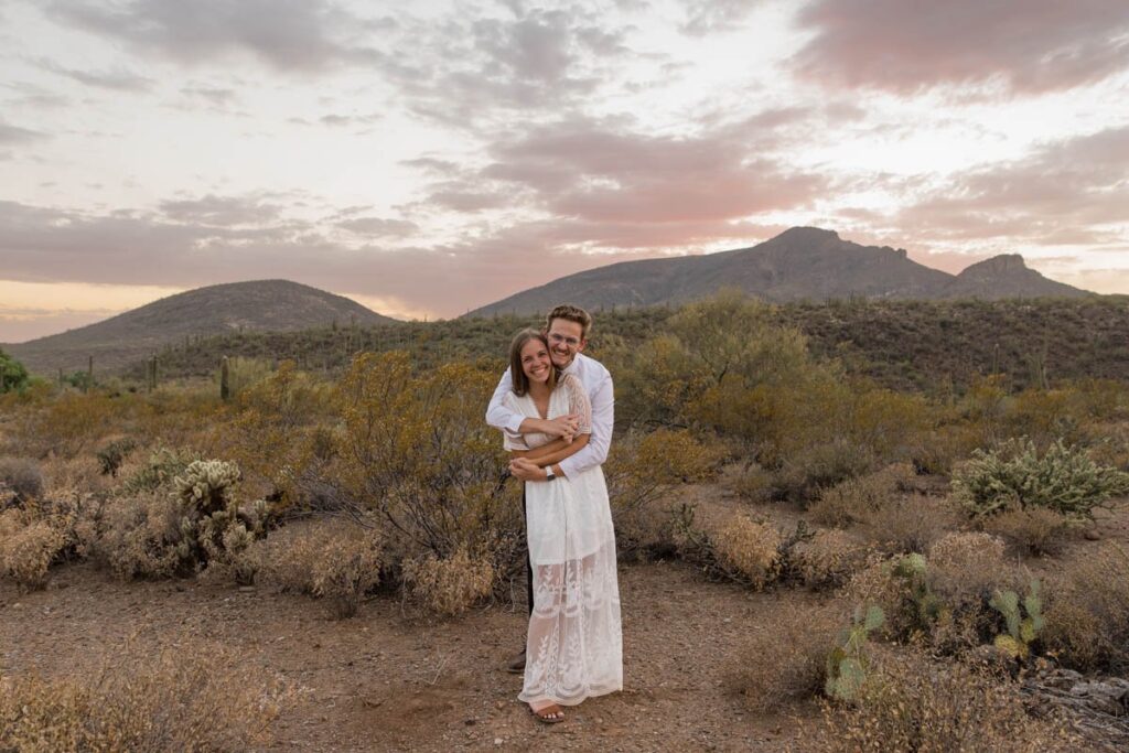 Groom wraps arms around bride as they stand in front of the mountains in the desert.
