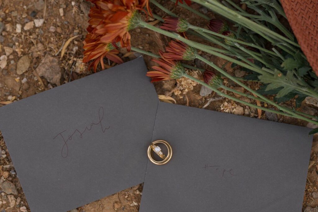 Wedding rings sit on grey envelopes next to rust colored flowers.