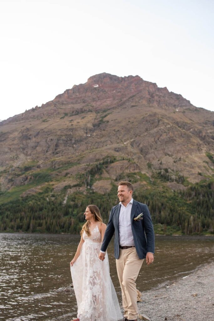 Couple walking along shore of Two Medicine Lake with mountain behind them at their Glacier National Park Elopement.