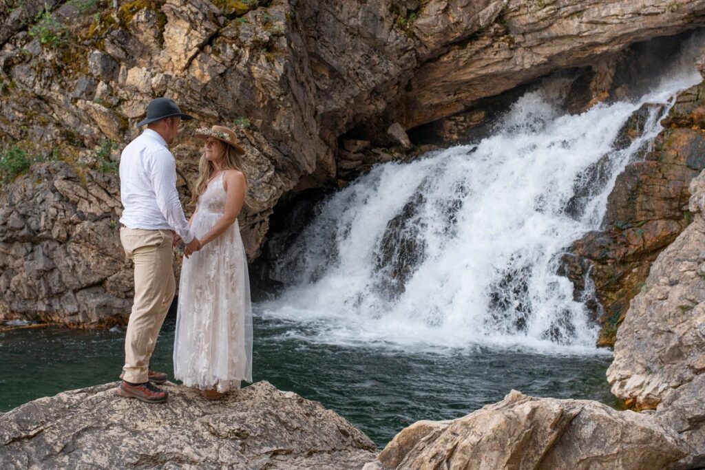 Couple stands in front of Running Eagle Falls on large rock holding hands.