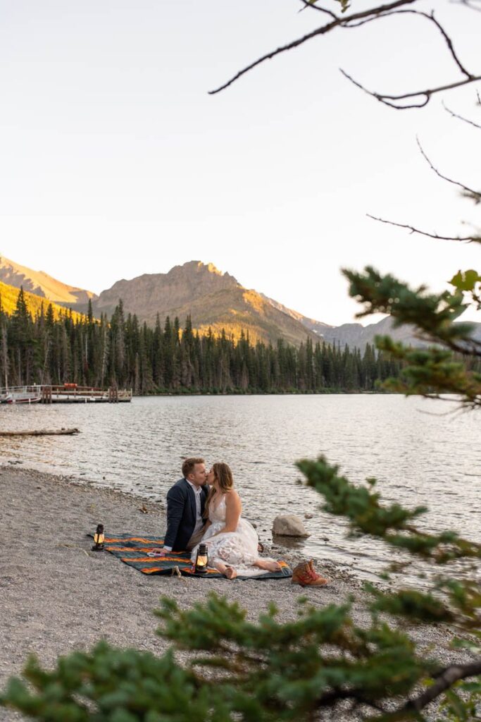 Couple kiss on a blanket in front of a lake with mountains in the background.