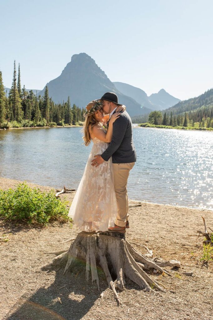 Couple stands together on tree stump in front of lake and mountains.