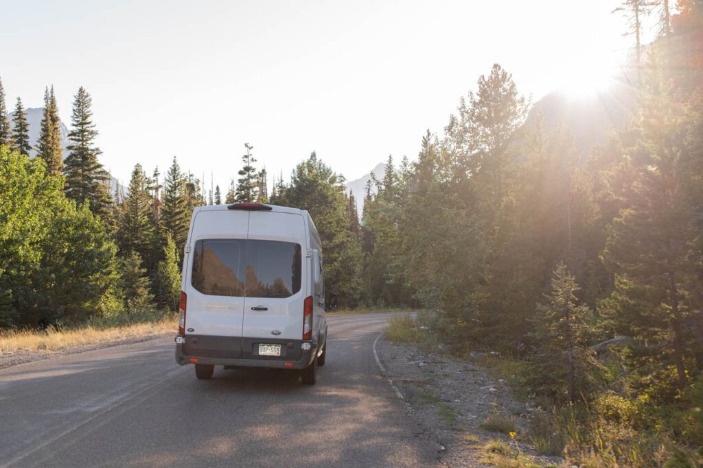 Couple driving camper van on mountain roads while sun sets behind mountains.