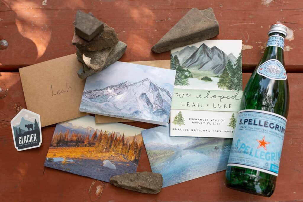 Casual wedding flatlay for a Glacier National Park Elopement with postcards, elopement announcement, stacked rocks, and sparkling water.