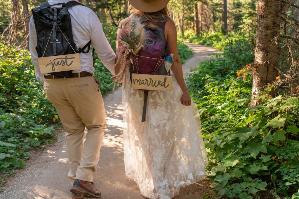 Couple hikes with backpacks that have "just married" signs handing from them during Glacier National Park Elopement.