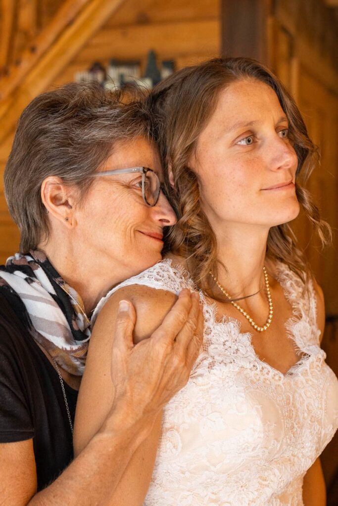 Mother stands behind bride hugging her after putting on pearl necklace.