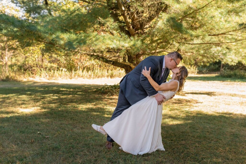 Groom dips bride and kisses her under evergreen trees.