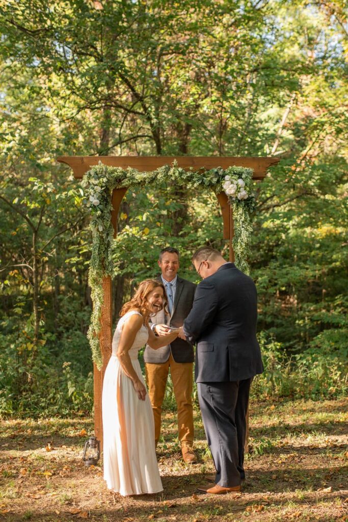 Bride, groom, and officiant laugh together during exchanging of rings.