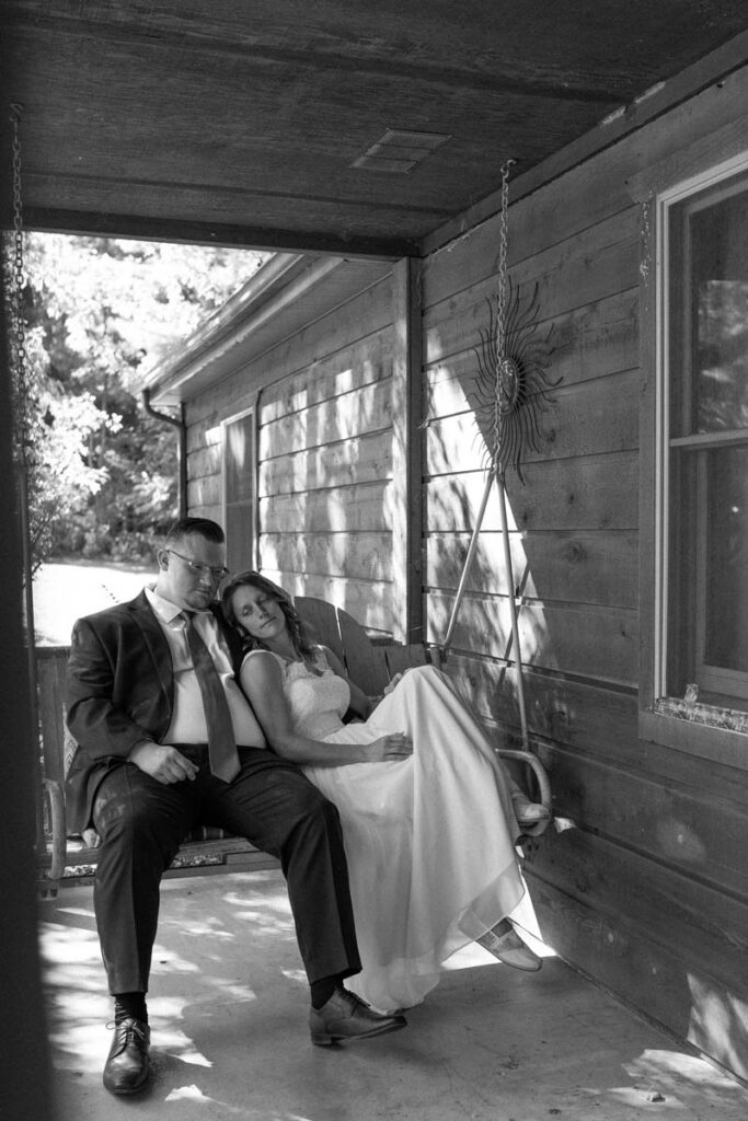 Couple sits quietly on porch swing with sun shining through trees casting shadows around them.