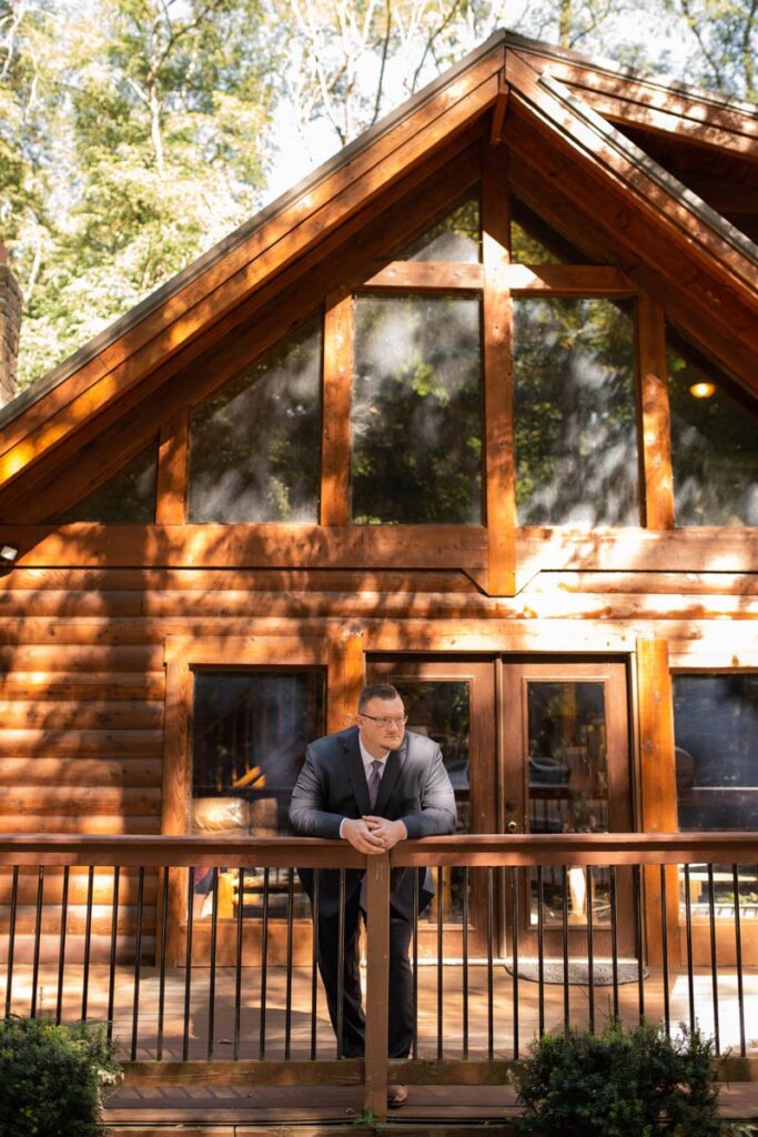 Groom leans on railing in front of log cabin while looking off into the distance.