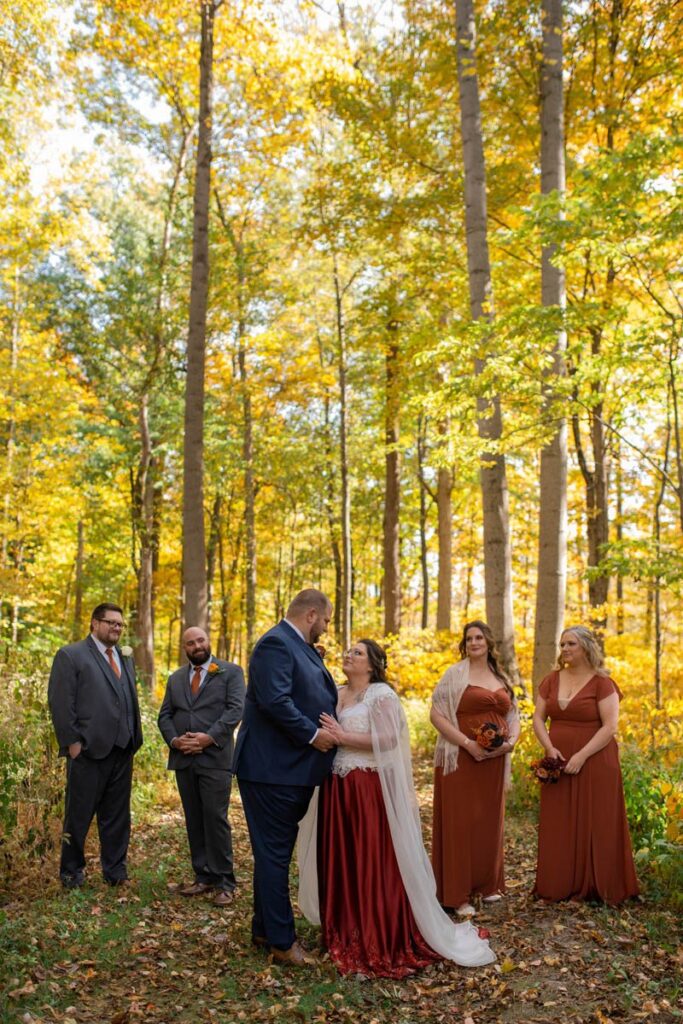 Bridal party smiles at bride and groom holding one another in the woods.