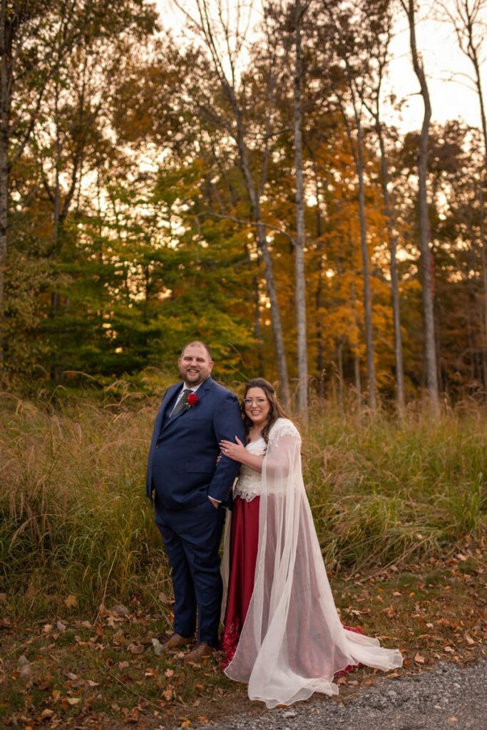 Bride and groom stand together in front of meadow and trees laughing while sun sets behind them in autumn.