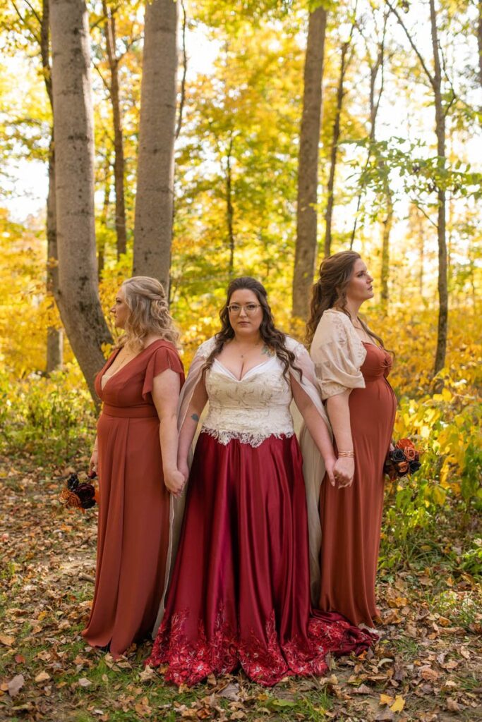 Bride holds hands with bridesmaids while standing in the woods together.