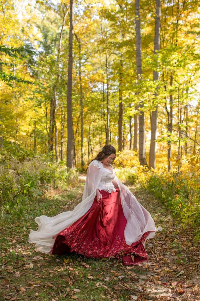 Bride twirls in her red wedding dress in the woods on a sunny, fall day.