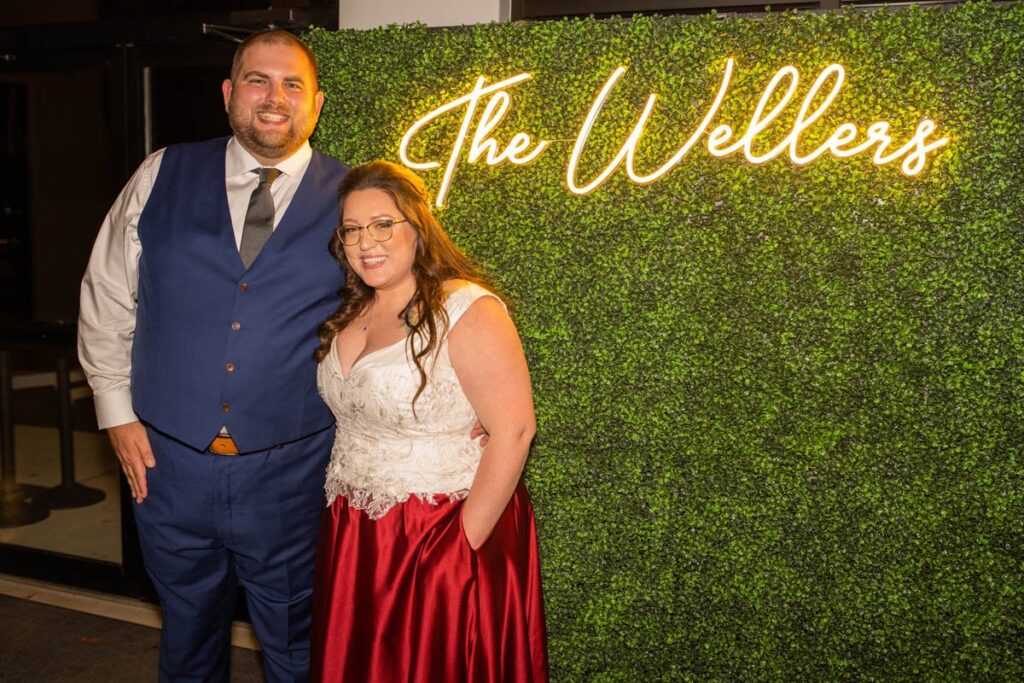 Bride and groom stand in front of custom neon sign of their last name at wedding reception.