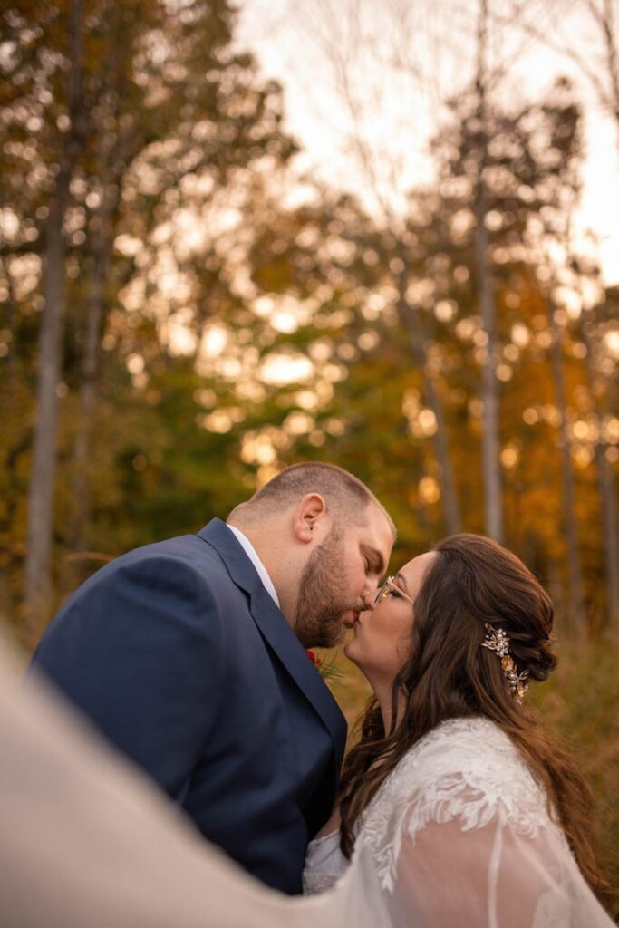 Bride and groom kiss with vibrant fall treeline behind them.