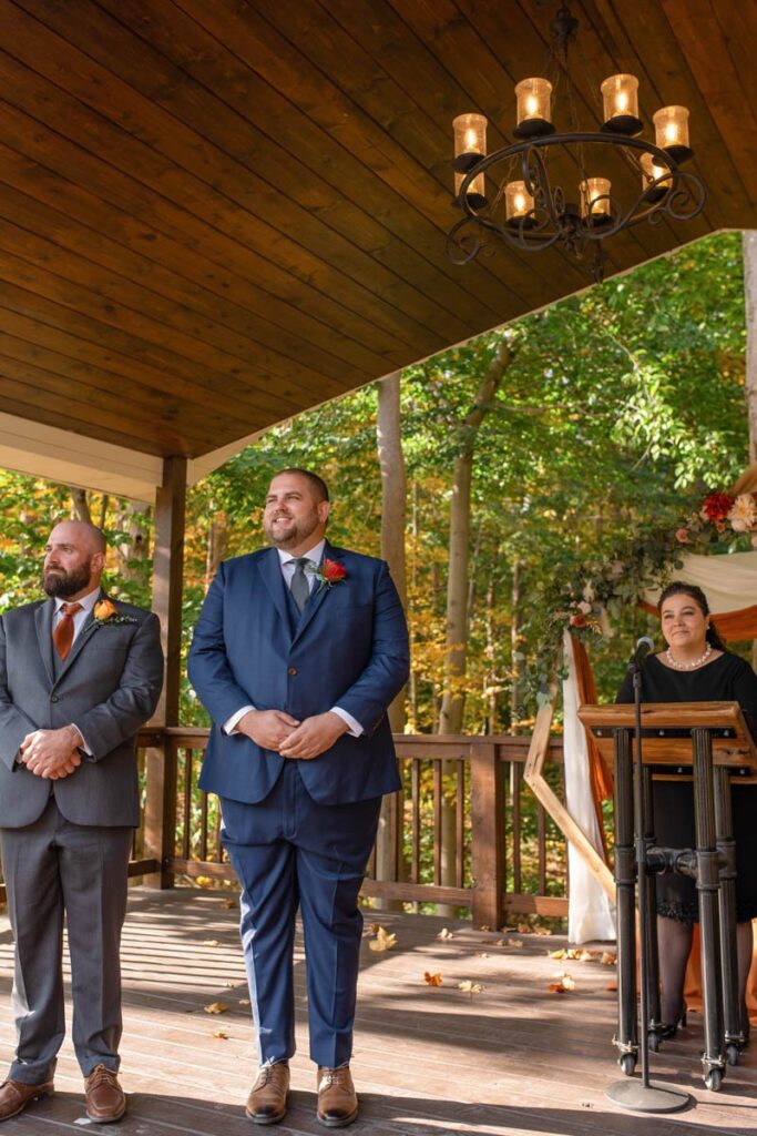 Groom smiles with best man and officiant while watching Bride walk down the aisle.