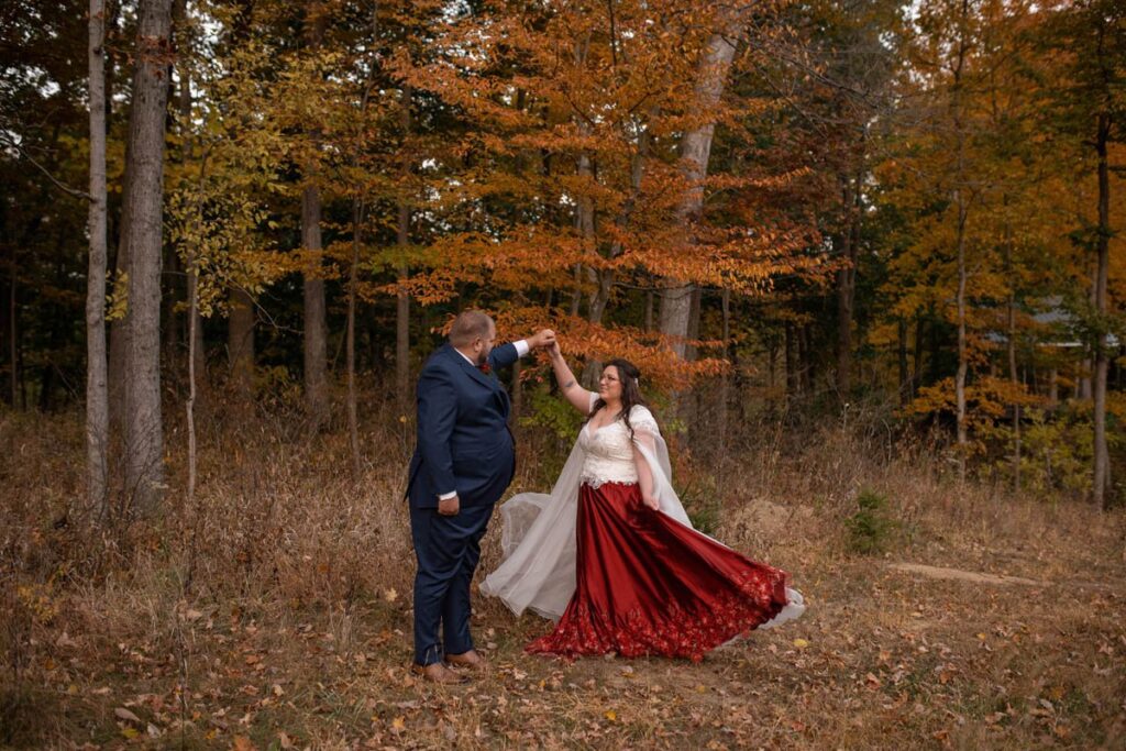 Groom twirls bride wearing a red dress in the woods during autumn.