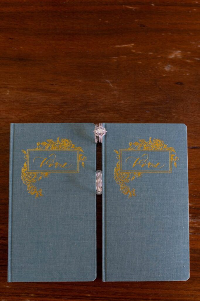Wedding rings sitting between two teal vow books on wooden table.