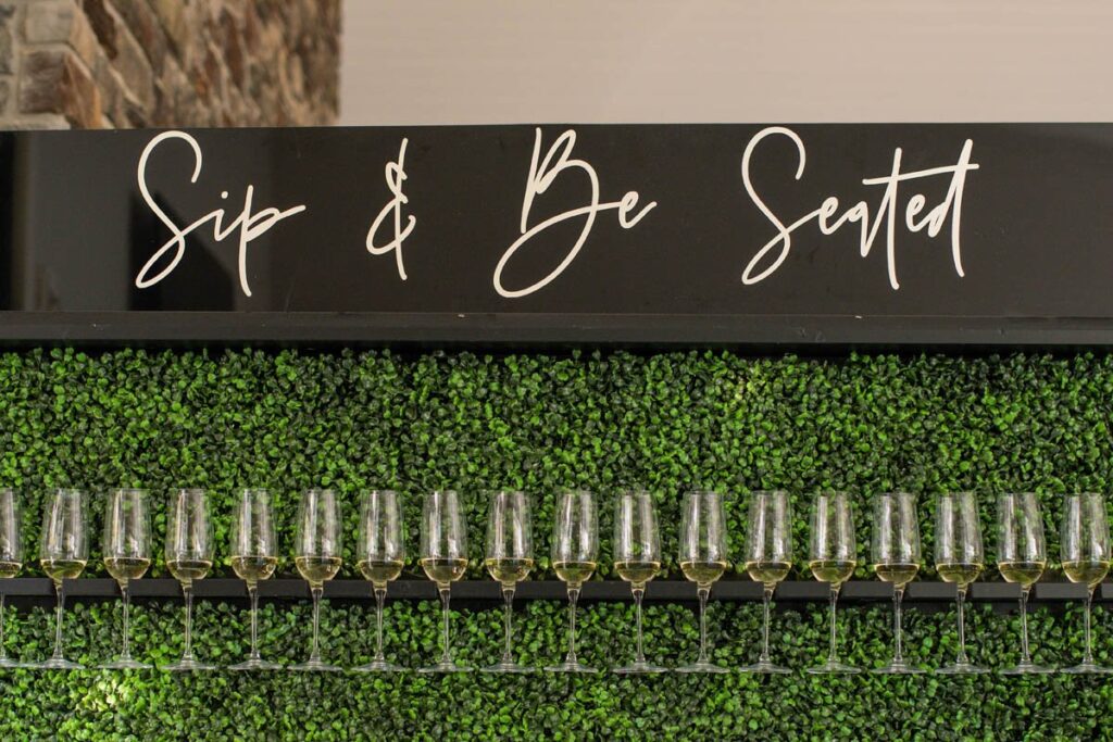 Sip and be seated champagne wall with greenery at Owl Ridge Wedding Venue.