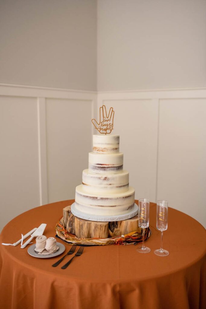 White wedding cake sits on log of wood with fall colored table cloth and "love long and prosper" wedding topper.