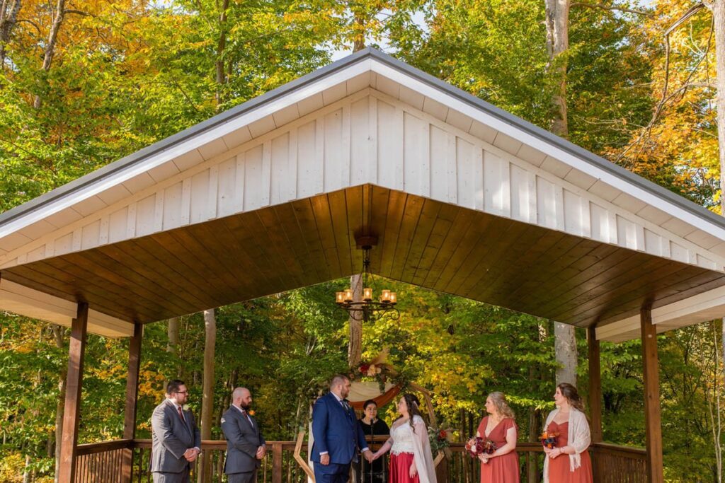 Bride and groom holding hands alongside bridal party and officiant during fall wedding ceremony.