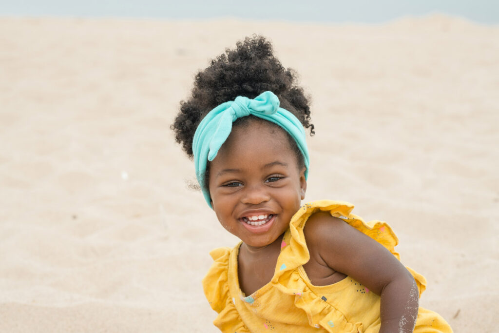 Little girl wearing teal headband smiles sitting in the sand.