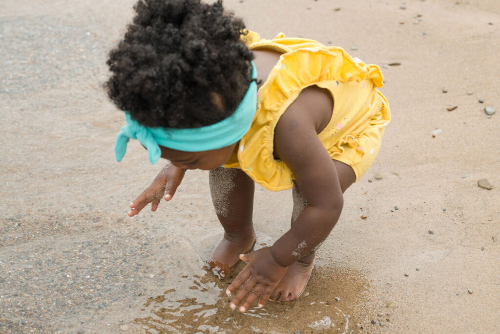 Little girl stands on shore and plays in sand and water.