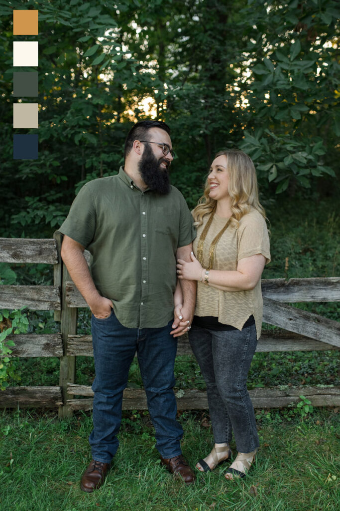 Couple looks at one another while standing in front of a wooded fence in the woods.