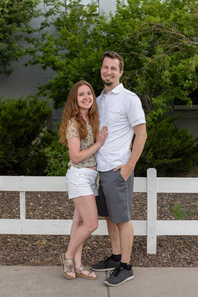Couple stands in front of white fence.