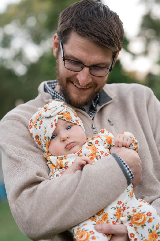Father smiles while holding his daughter who is wearing a yellow floral pattern outfit.