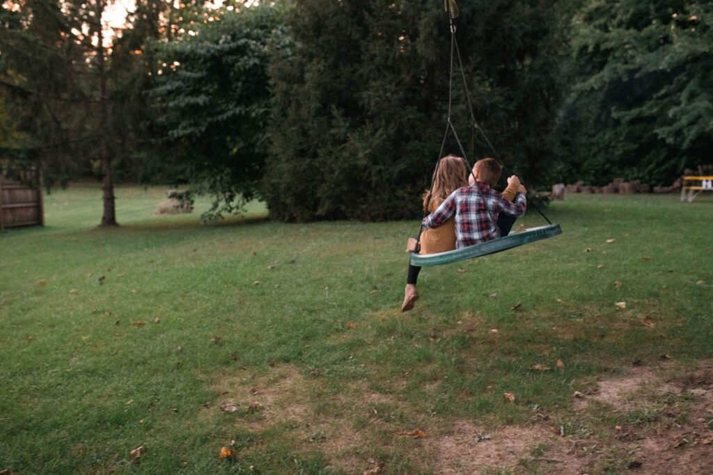 Brother has arm around sister while they swing together in their backyard.